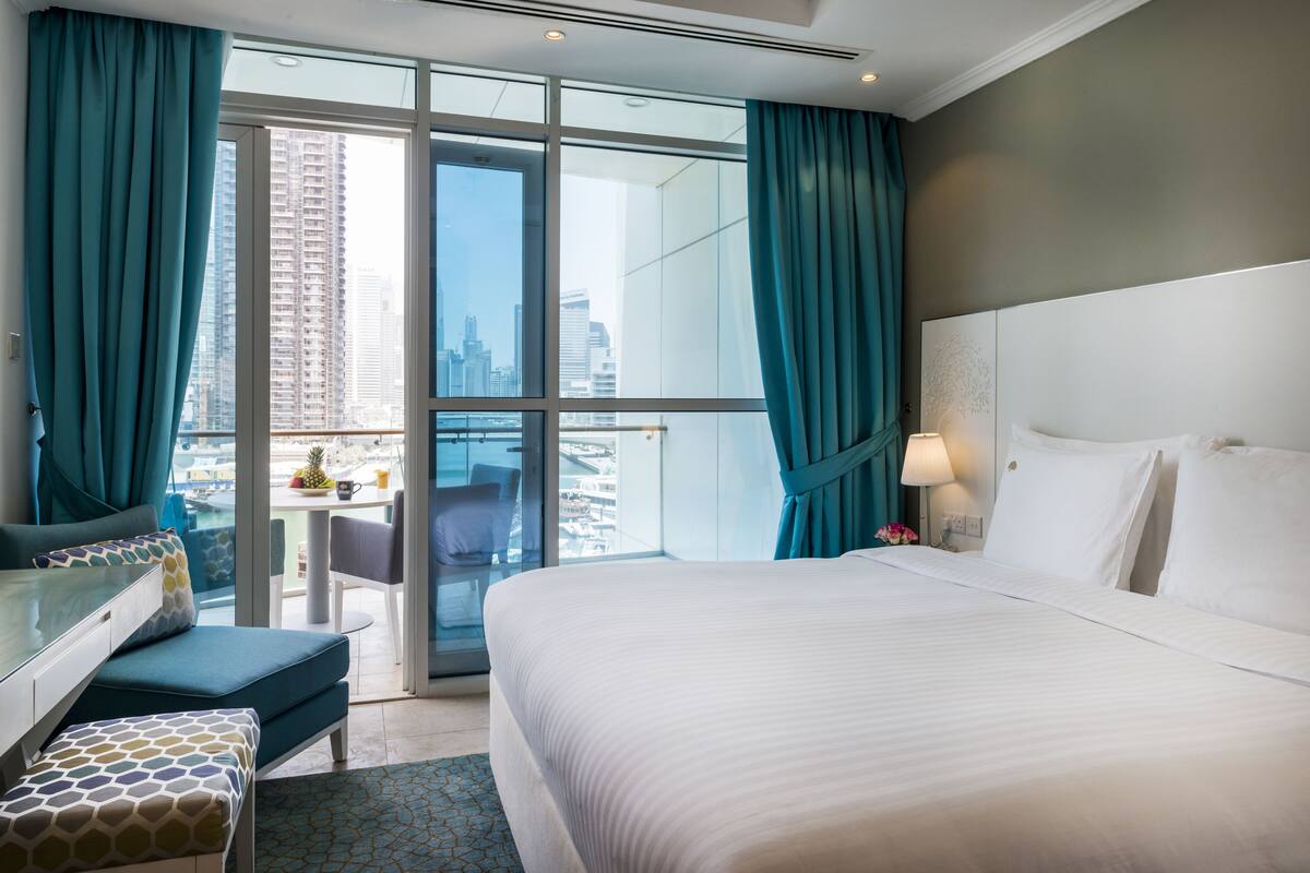 Habitación de hotel con una cama grande, un escritorio con una silla, una mesita con otra silla y vistas a la ciudad a través de amplios ventanales.