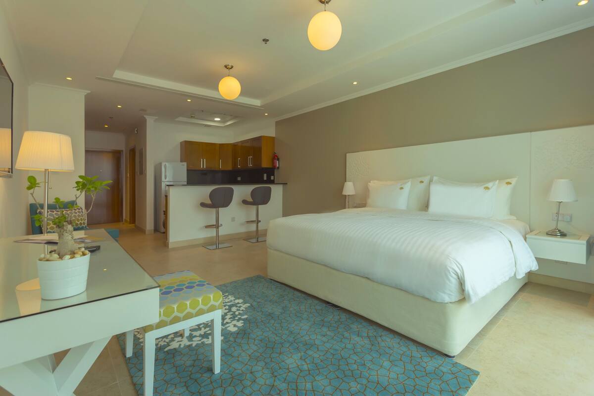 Una habitación de hotel moderna con una cama grande, un escritorio con lámpara, una pequeña cocina y una alfombra azul estampada.
