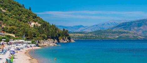 Una playa con aguas azules cristalinas, un muelle de madera y montañas al fondo.