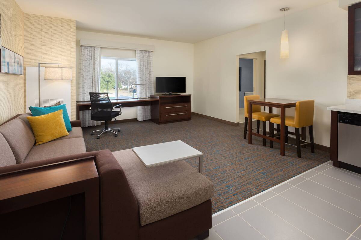 A modern hotel room with a sofa, a small table, a desk with a chair, a TV, a dining area with chairs, and a kitchen area.