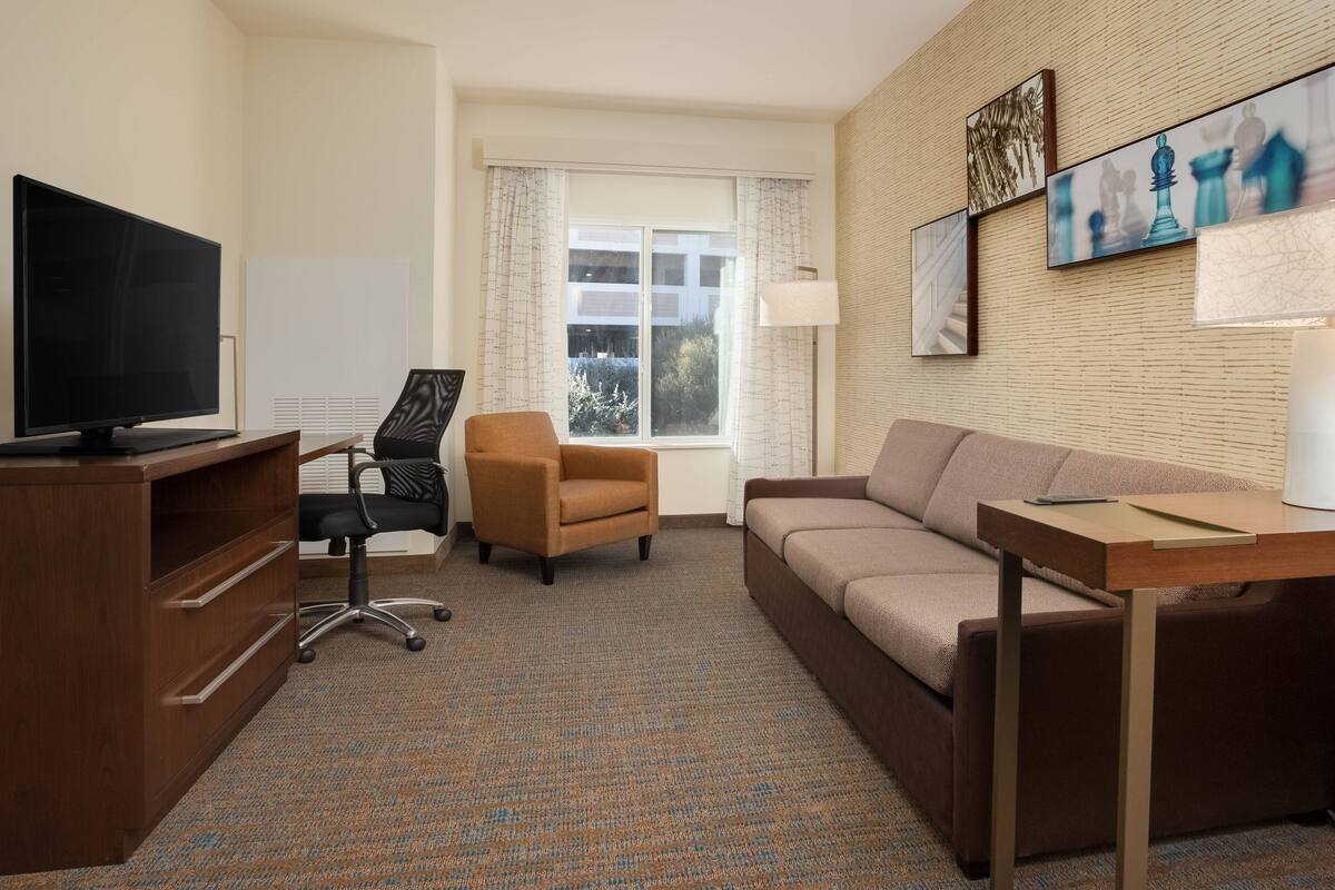 A hotel room with a flat-screen TV, a wooden desk, a brown armchair, a beige sofa, and a large window with curtains.