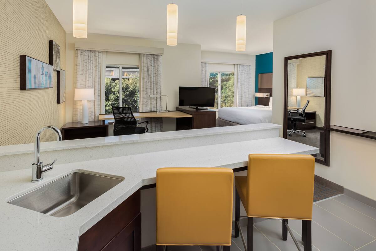 A modern hotel room with a kitchen area, a bed, a desk, and a large mirror.
