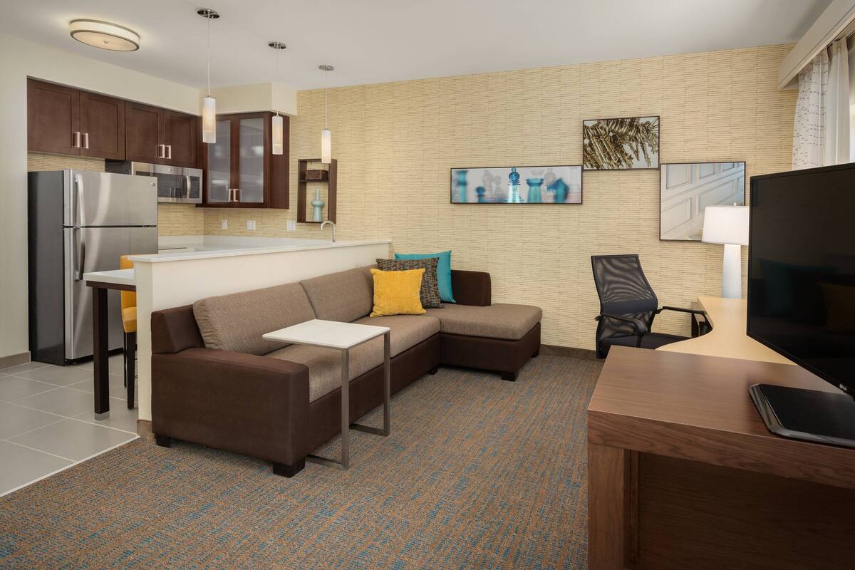 A modern living room with a sectional sofa, a flat-screen TV, a small table, and a kitchen area with a refrigerator and microwave.