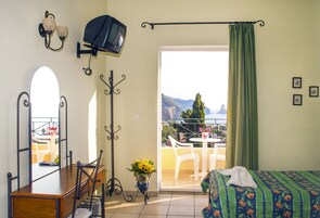 Habitación de hotel con balcón, cama con cubrecama floral, escritorio con silla, espejo, televisor y florero con flores.