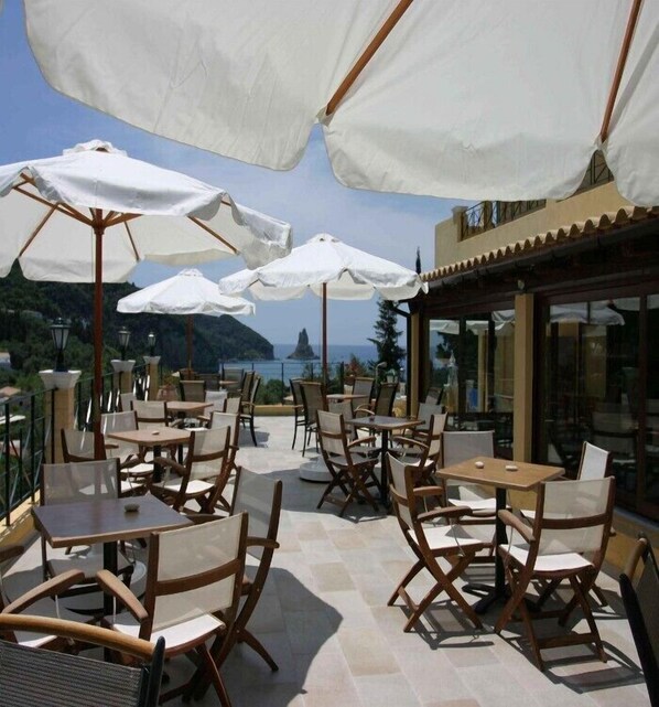 An outdoor patio with tables and chairs, white umbrellas, and a view of the sea and hills. - Alonakia Hotel (Corfu)