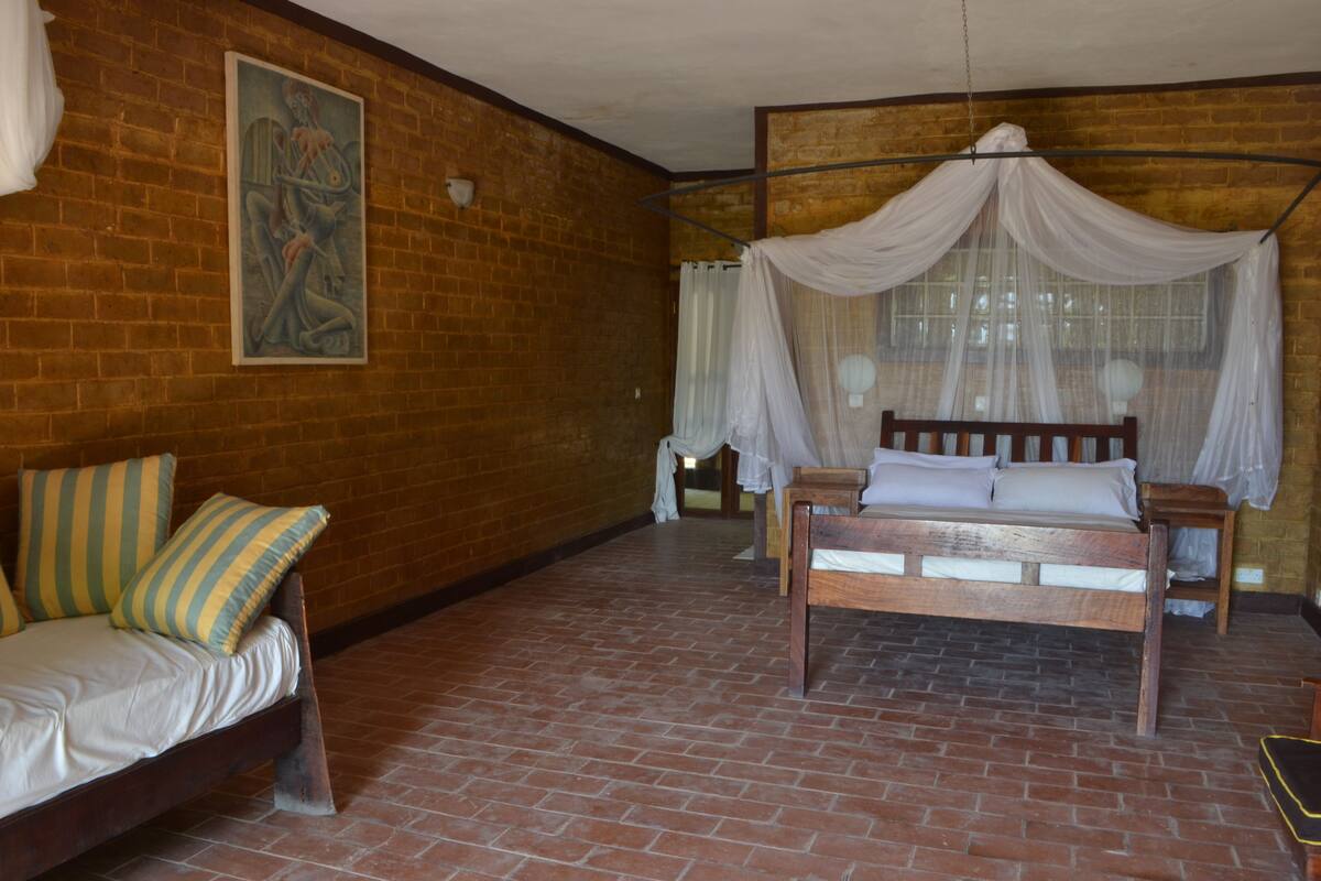 A room with a brick floor, a wooden bed with a canopy, a wooden bench, and a striped cushion.