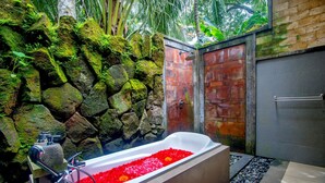 A modern bathroom with a stone wall, a wooden door, and a bathtub filled with red flowers. - Alam Dania Cottage (Ubud)