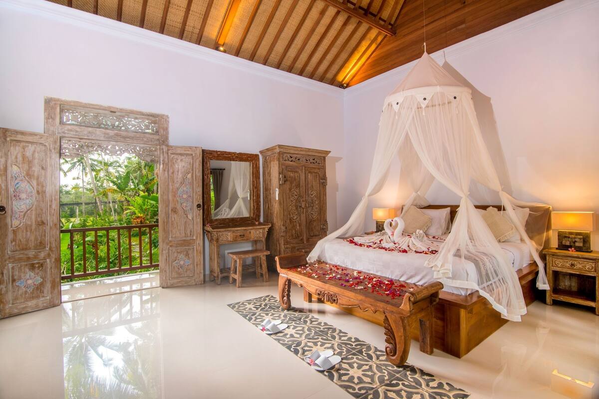 Quarto com cama de dossel, móveis de madeira, um espelho e vista para o verde lá fora.