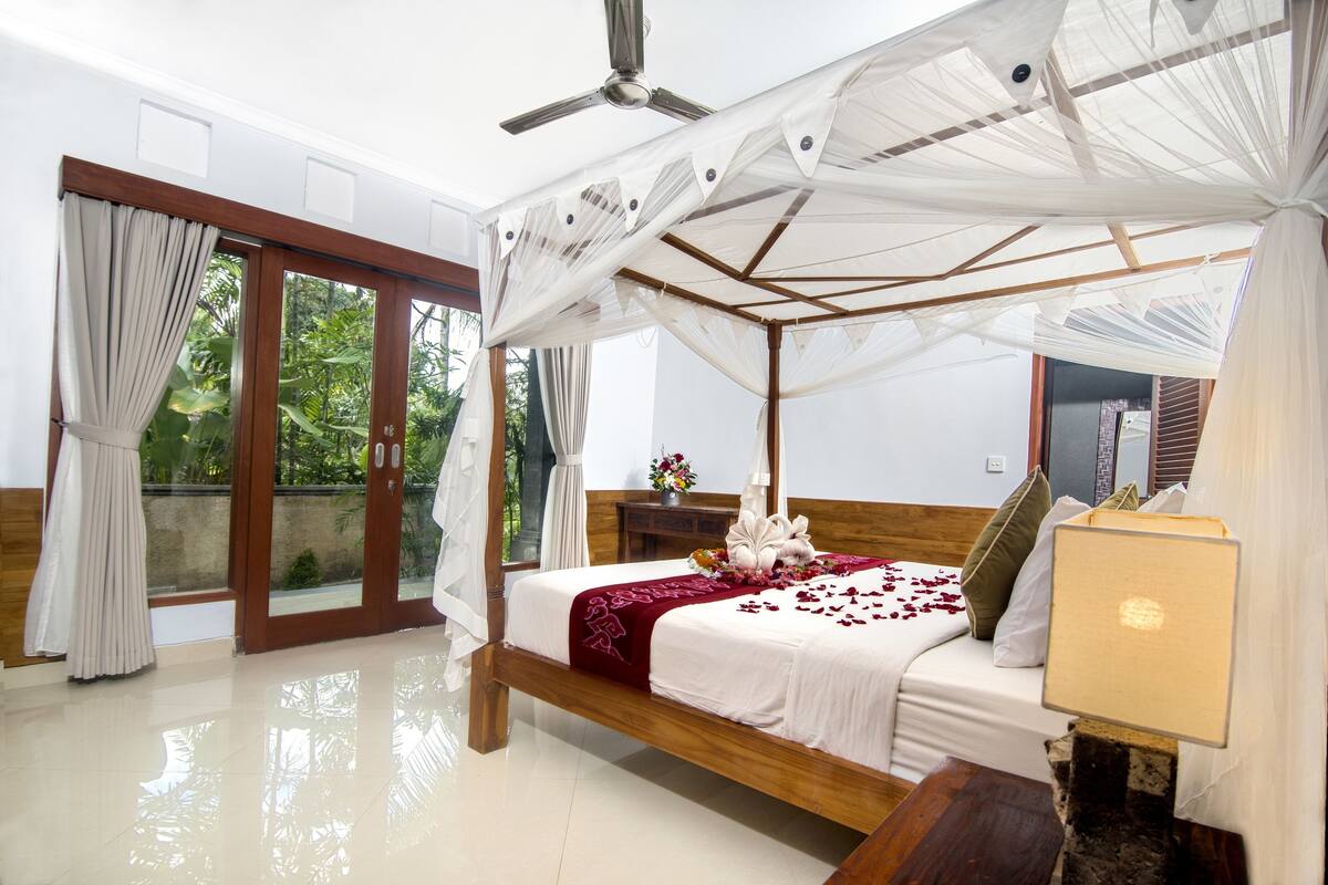 Quarto com cama de dossel, cabeceira de madeira, mesa de cabeceira com abajur e vista para o verde através de uma porta de vidro deslizante.