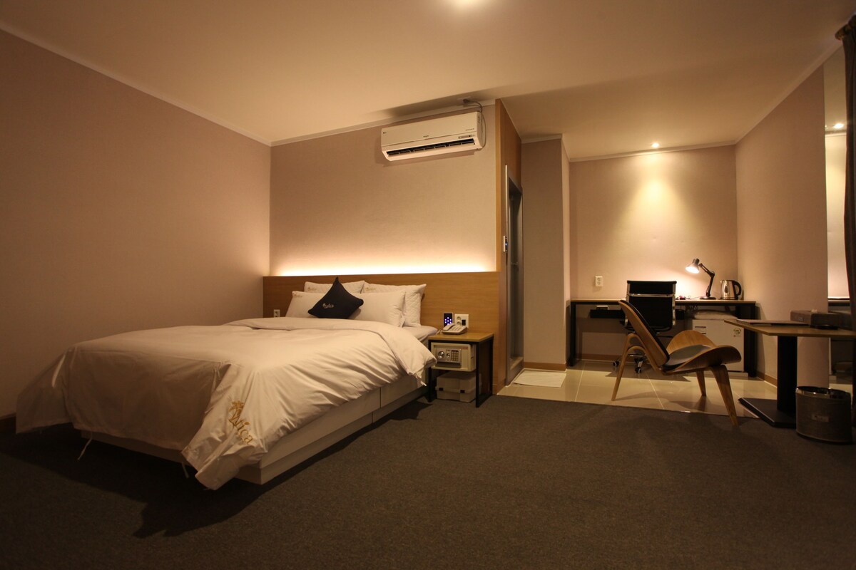 A hotel room with a bed, bedside table, lamp, chair, desk, and an air conditioning unit.