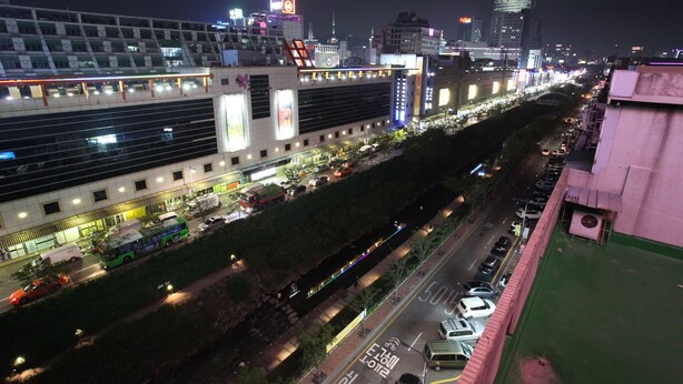 A nighttime cityscape with illuminated buildings, a busy street, and a green space with a pathway. - Vatica Hotel Dongdaemun (Seoul)