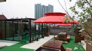 An outdoor patio area with wooden tables and red umbrellas, surrounded by glass walls and green artificial turf. - Lombard House - Hostel (Seoul)