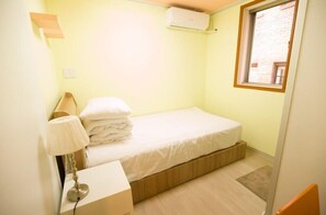 A small, single-bed room with a wooden platform bed, a bedside lamp, a wall-mounted air conditioner, and a window with a view of a brick building.
