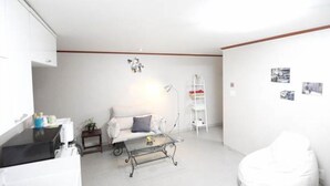 A modern living room with a white sofa, a glass coffee table, and a white armchair. There is a small side table with a lamp and a few decorative items. - Lombard House - Hostel (Seoul)