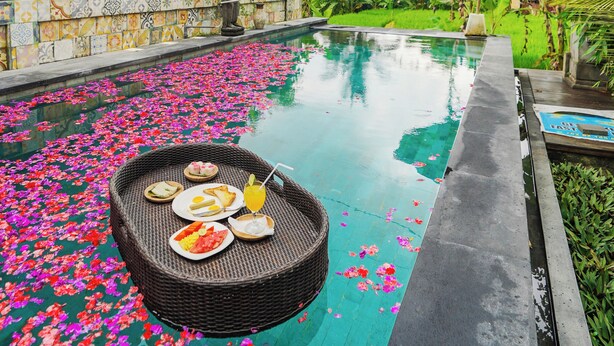 A pool with floating flower petals, a wicker table with plates and drinks, and a tiled wall with a decorative pattern.