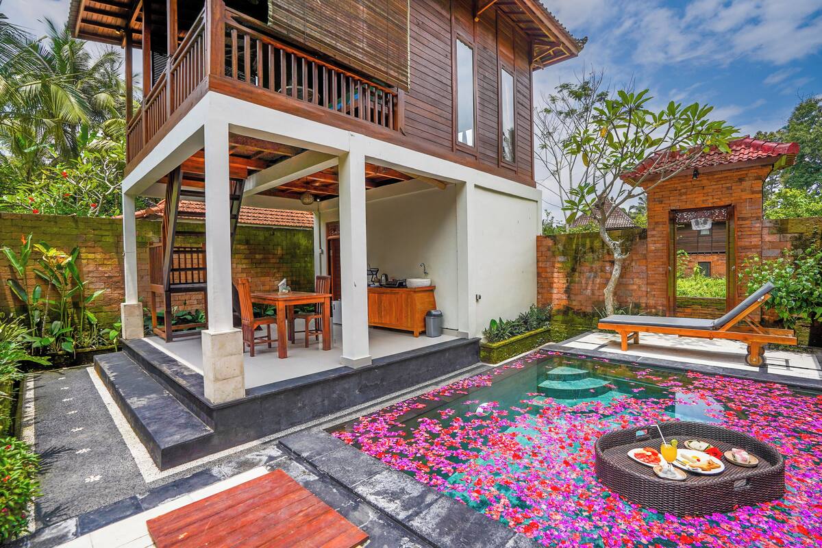 A two-story house with a balcony, a swimming pool surrounded by flower petals, and a dining area with a table and chairs.