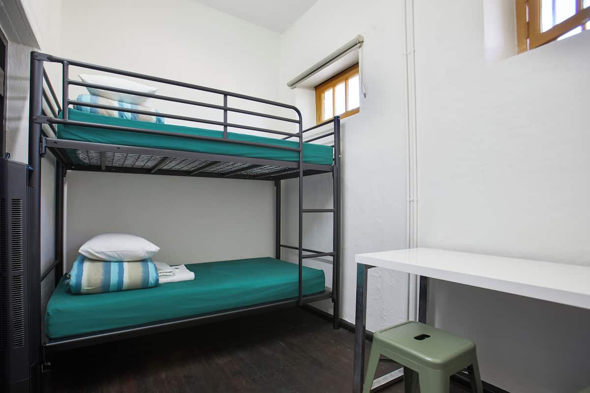 Un lit superposé avec un matelas vert, un oreiller blanc et une couverture à rayures. On y trouve un petit tabouret vert et un bureau blanc, avec une fenêtre au-dessus.
