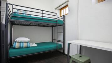 Een stapelbed met een groene matras, een wit kussen en een gestreept dekbed. Er staat een kleine groene kruk en een wit bureau met daarboven een raam.
