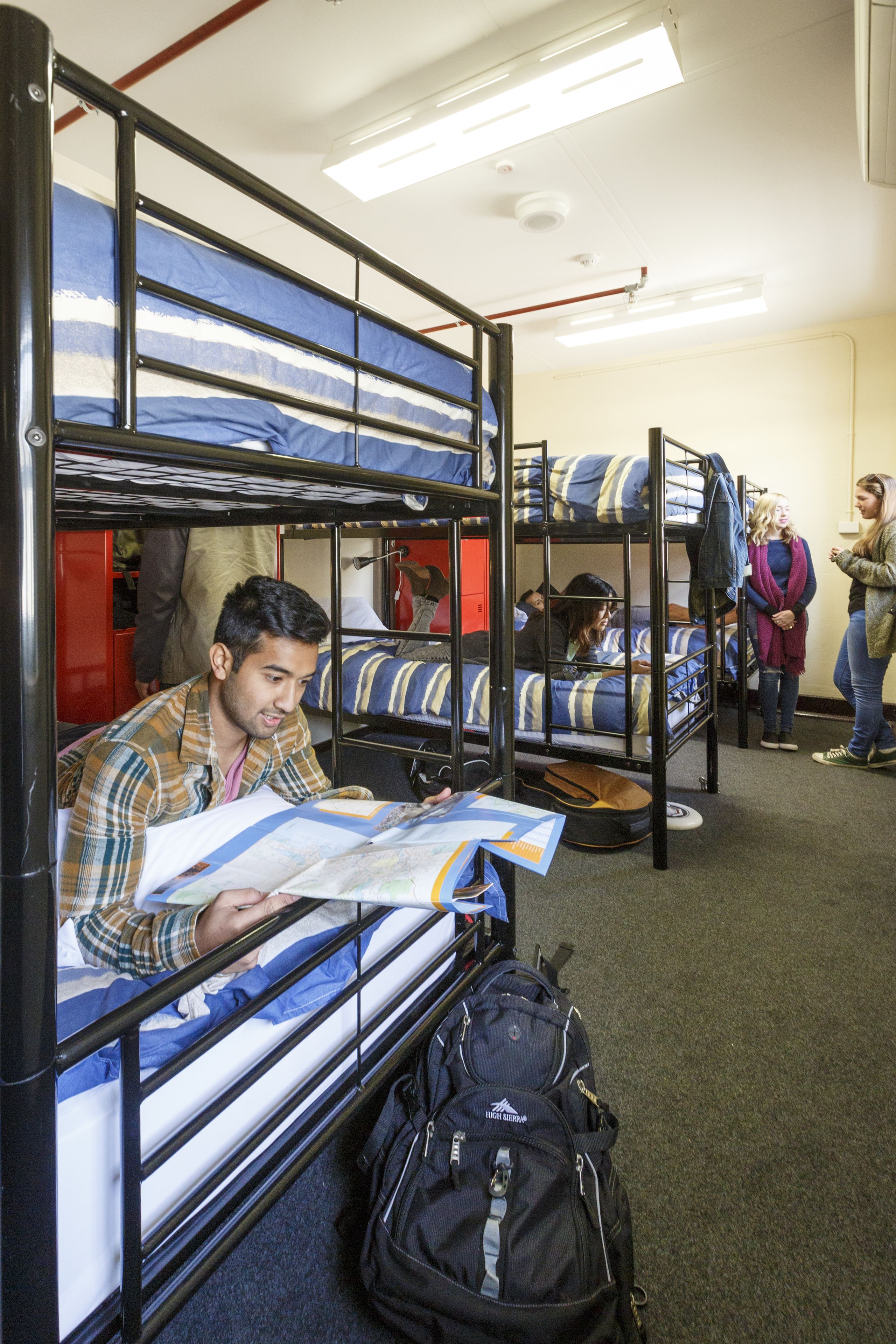 A dormitory room with bunk beds, a black backpack on the floor, and a person reading a book.