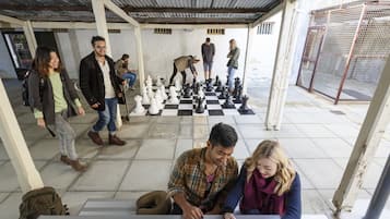 Un grupo de personas jugando ajedrez en un espacio al aire libre cubierto, con suelo a cuadros y valla metálica.