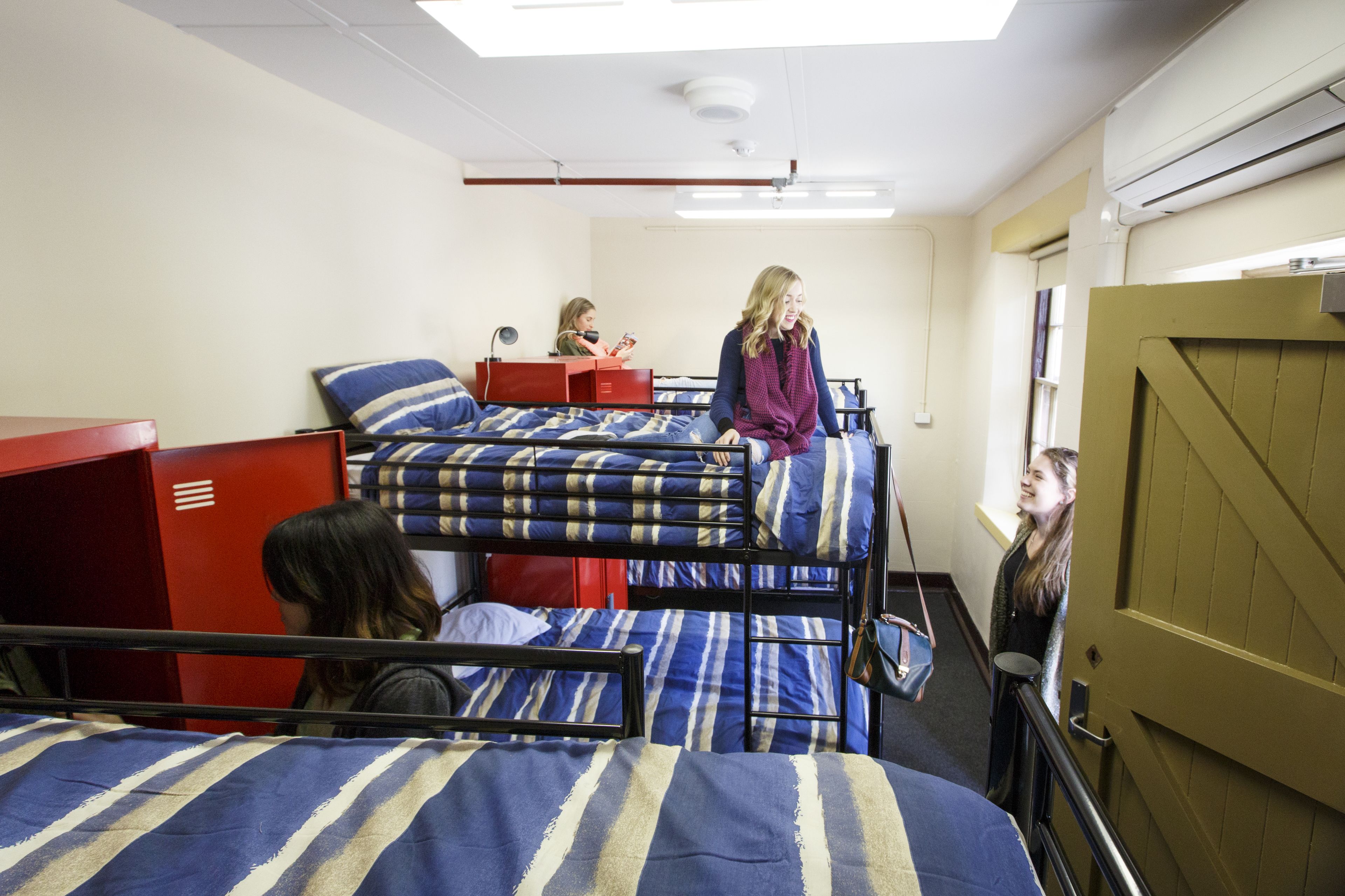 A dormitory room with bunk beds, a red storage unit, and a window with curtains.