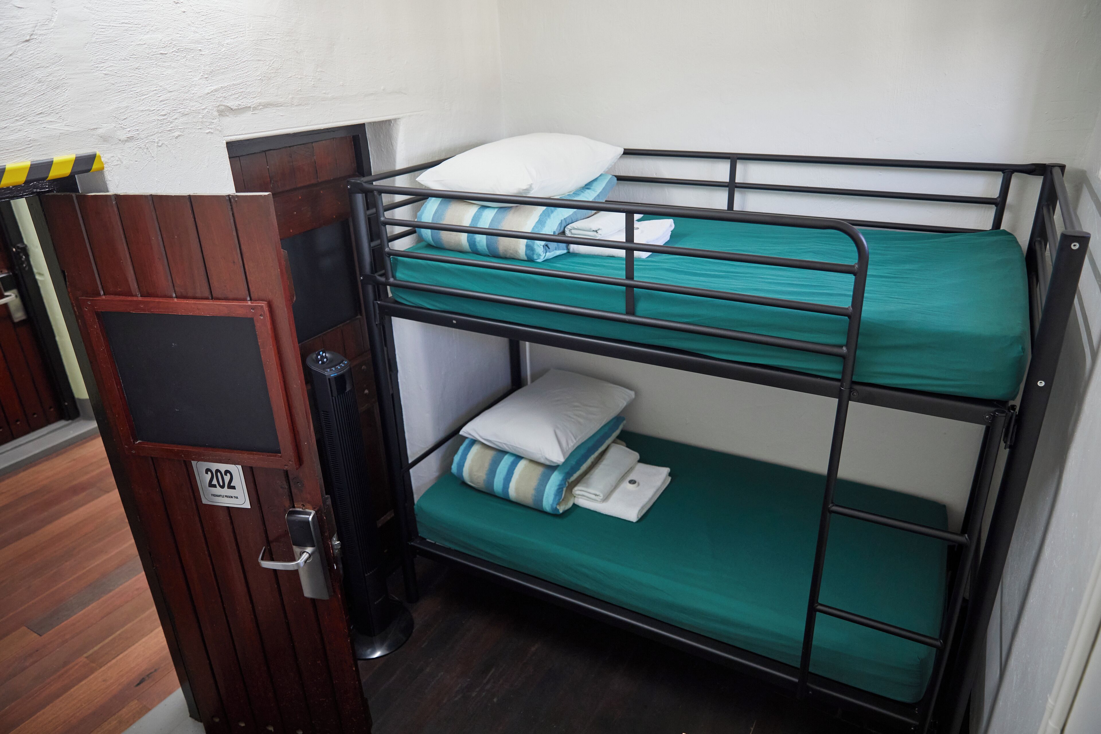 Cama litera con dos niveles, cada uno con colchón y almohadas verdes. Se ve una puerta con placa de número.