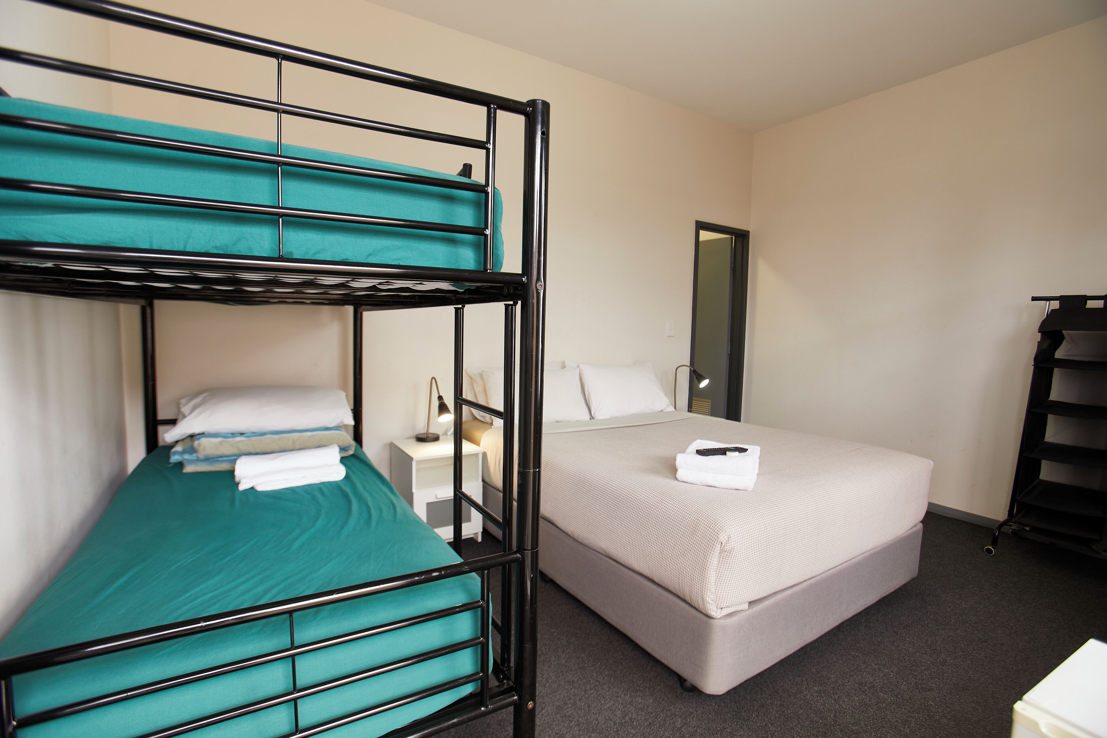 A hotel room with a bunk bed, a single bed, a bedside table with a lamp, and a mirror on the wall.