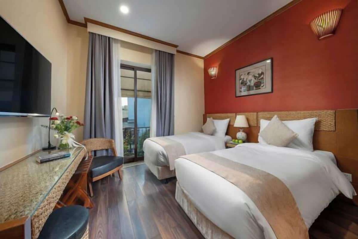 Habitación de hotel con dos camas, un escritorio con silla, televisión y balcón con cortinas.