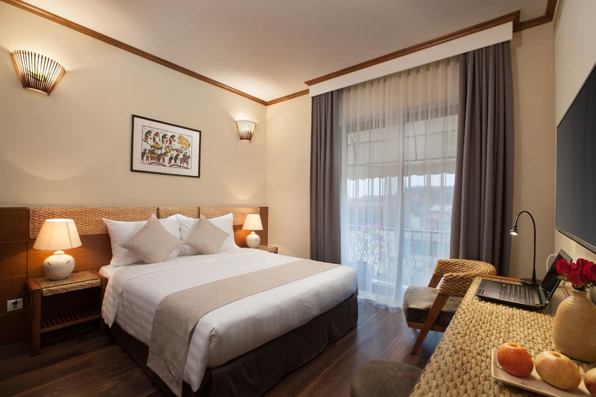 Una habitación de hotel con una cama grande, lámparas de noche, un escritorio con una computadora portátil, un florero con flores y un cuadro en la pared.