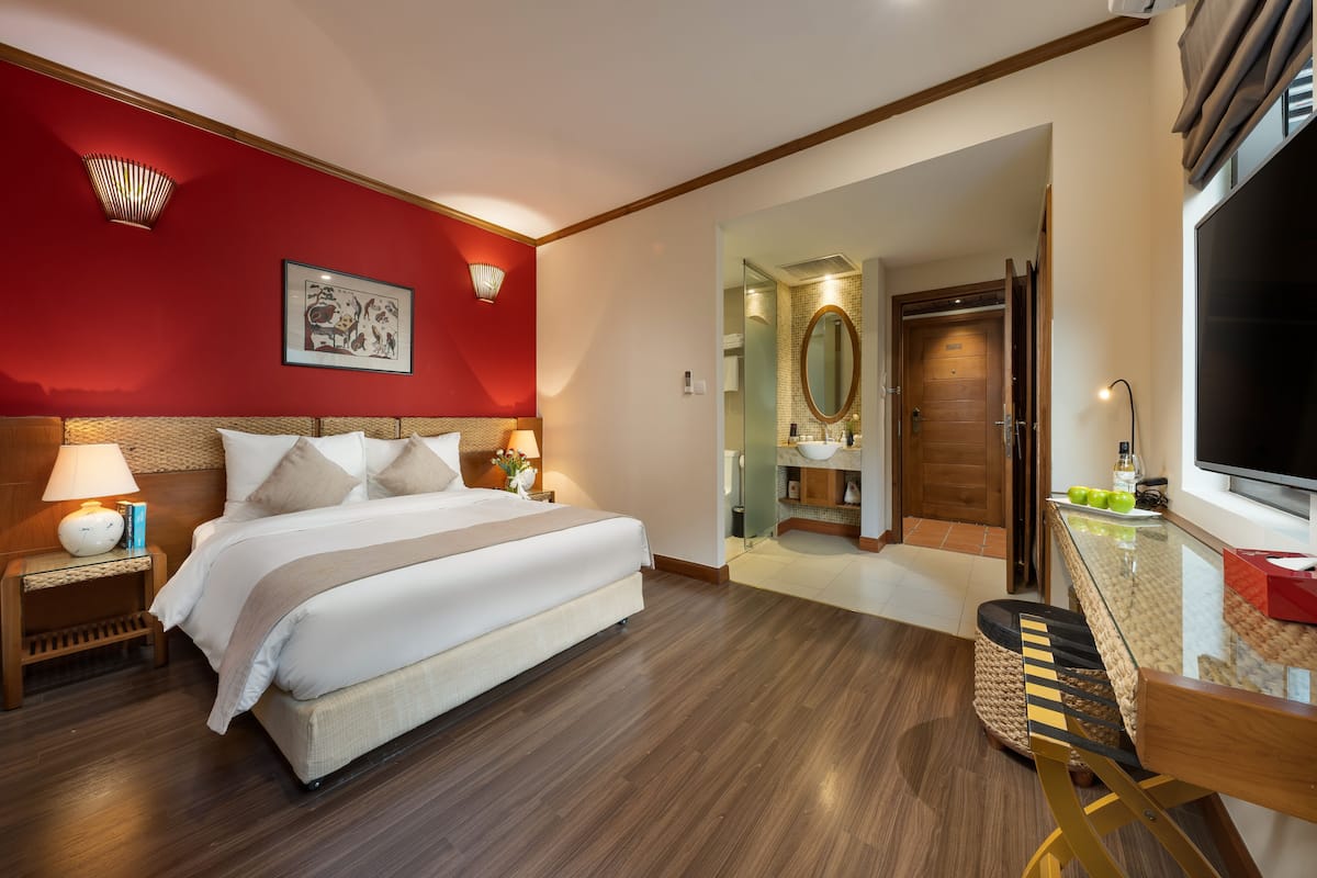 Una habitación de hotel con una cama grande, lámparas de noche, una pared de acento roja, suelo de madera, una mesa de cristal con una silla y un televisor.