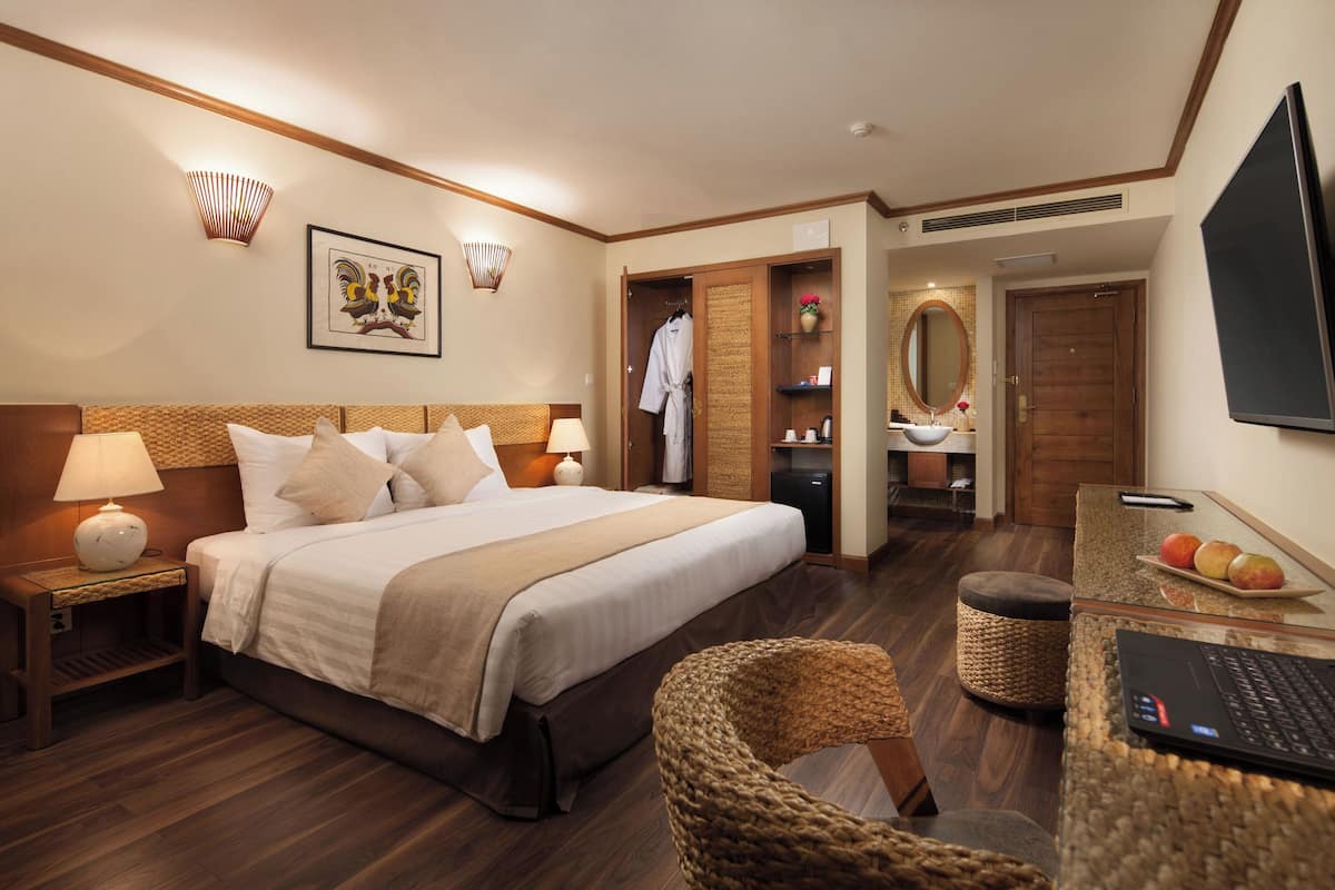 Habitación de hotel con cama, mesitas de noche, un escritorio con una computadora portátil, una silla, una pequeña mesa con fruta, un espejo y un armario.