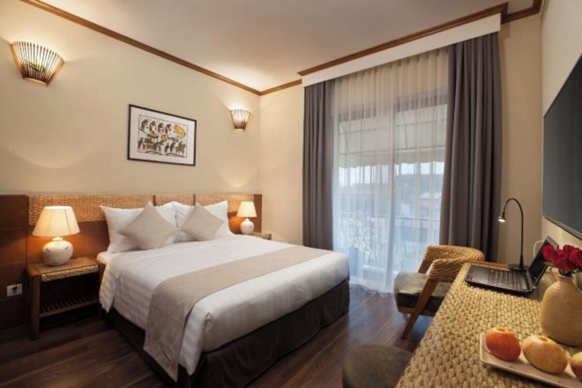 A hotel room with a large bed, two bedside lamps, a desk with a laptop, a vase with flowers, and a large window with curtains.