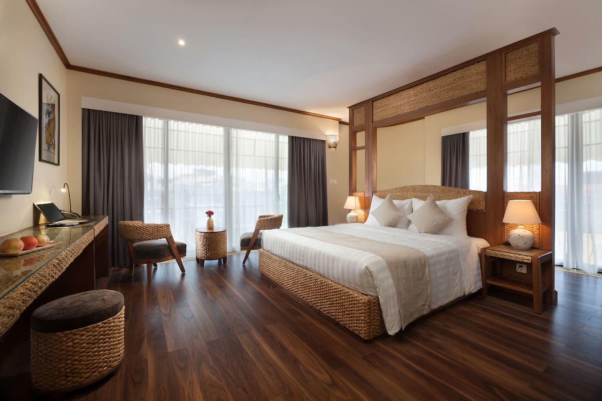Amplia habitación de hotel con una cama grande, un escritorio con una computadora portátil, una silla y una mesita de noche con una lámpara.