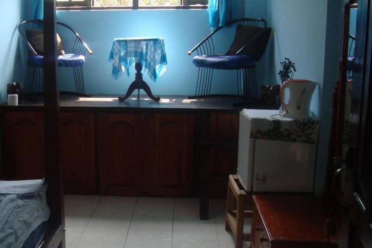 A small, dimly lit room with a bed, a wooden cabinet, a table with a blue tablecloth, and two chairs.