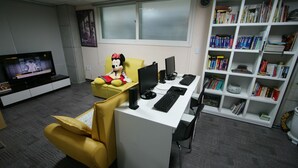 Una oficina moderna con un escritorio, una computadora y un peluche. Se ve una estantería con libros y un televisor al fondo.
