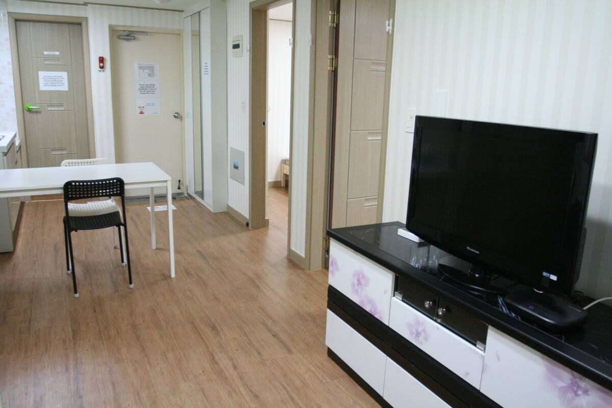A hotel room with a flat-screen TV, a dining table with chairs, and a kitchenette area.