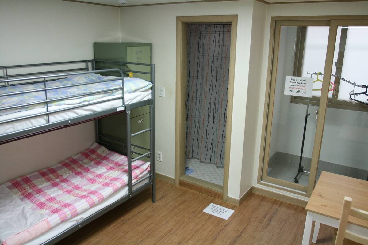 A dormitory room with bunk beds, a small table, and a door leading to a bathroom.