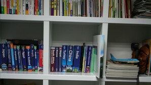 Una estantería con libros de diversos temas, una pequeña maqueta de un edificio y un cuenco decorativo.