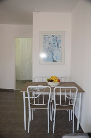 Un coin repas avec une table et deux chaises blanches, un saladier de fruits sur la table et un tableau abstrait encadré au mur.