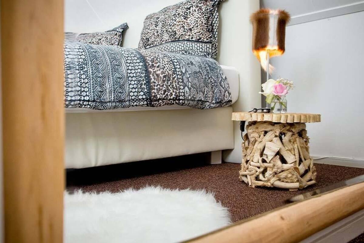 A cozy living room with a white sofa, patterned cushions, a small wooden side table with a vase of flowers, and a fluffy white rug.