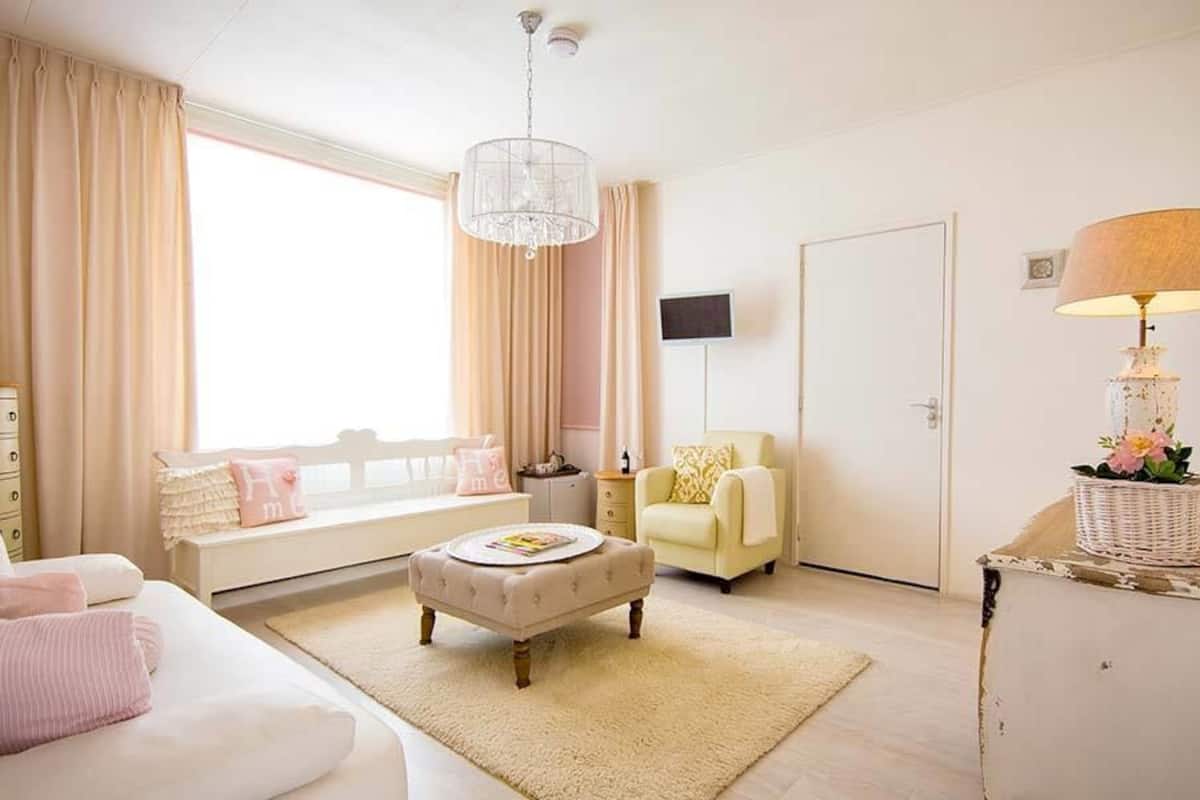 A well-lit bedroom with a bed, a sitting area with a chair and ottoman, a dresser, a television, and a lamp.