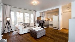 Una sala de estar moderna con un sofá blanco, un reposapiés gris y una lámpara de pie. Se aprecia una cocina al fondo con armarios blancos y una campana extractora de acero inoxidable.