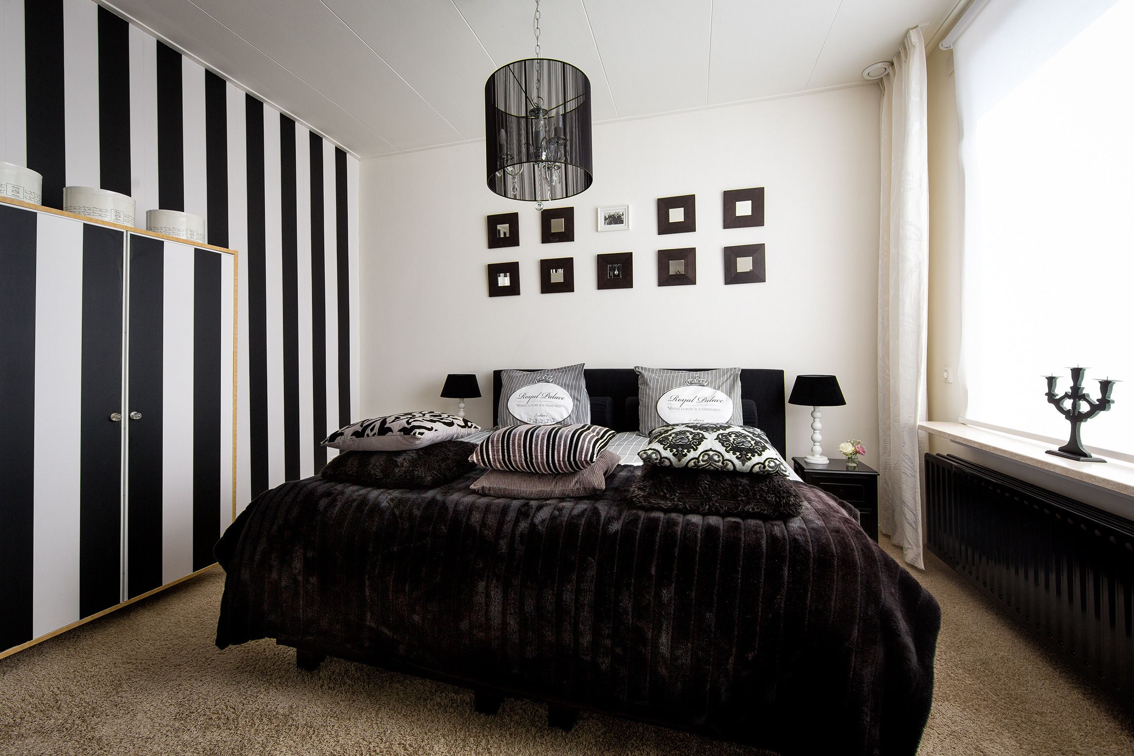 Ein Schlafzimmer mit einem schwarz-weiß gestreiften Kleiderschrank, einem Bett mit vielen Kissen, einer Chandelüster, gerahmten Bildern an der Wand und einem Fenster mit einem Kerzenleuchter.