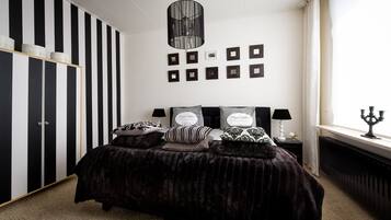 Ein Schlafzimmer mit einem schwarz-weiß gestreiften Kleiderschrank, einem Bett mit vielen Kissen, einer Chandelüster, gerahmten Bildern an der Wand und einem Fenster mit einem Kerzenleuchter.