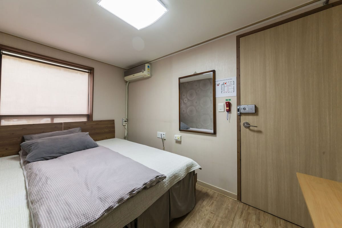 Une chambre à coucher comprenant un lit, une fenêtre avec des stores, un appareil de climatisation, une porte et un interrupteur fixé au mur.