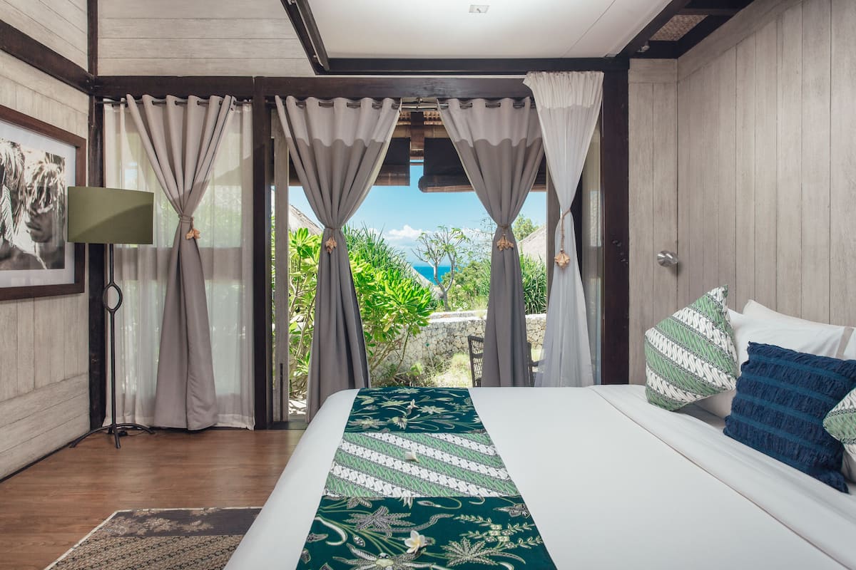 A bedroom with a large bed, curtains, a view of greenery, and a mounted television.