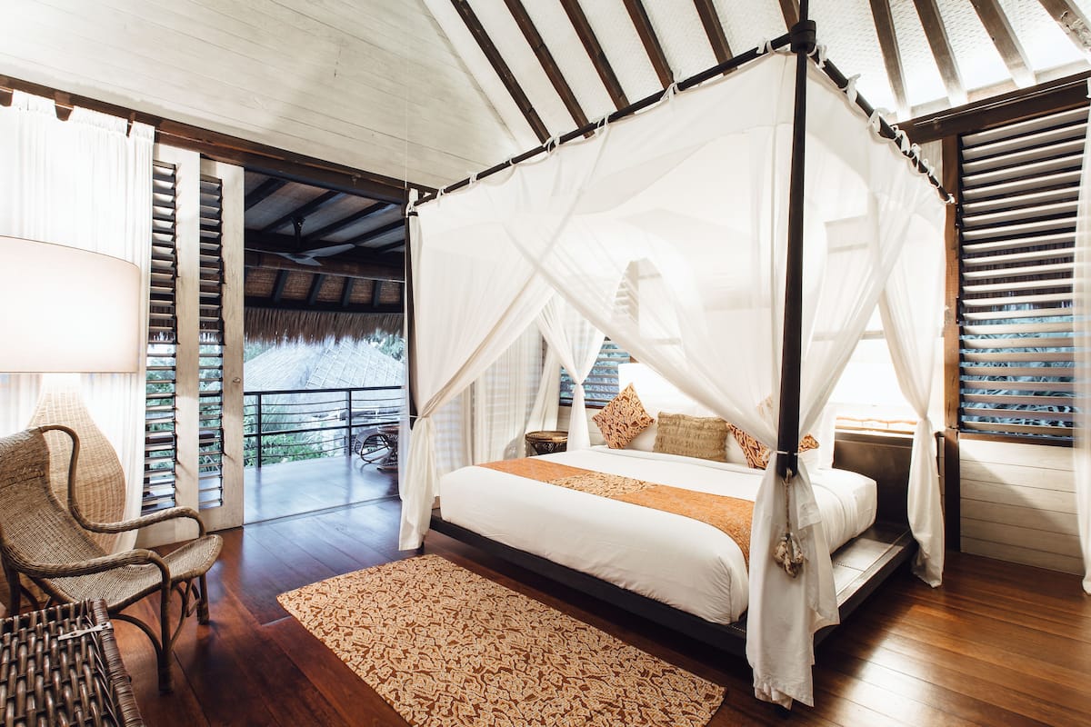 A bedroom with a four-poster bed, a wicker chair, and a large window with a view.