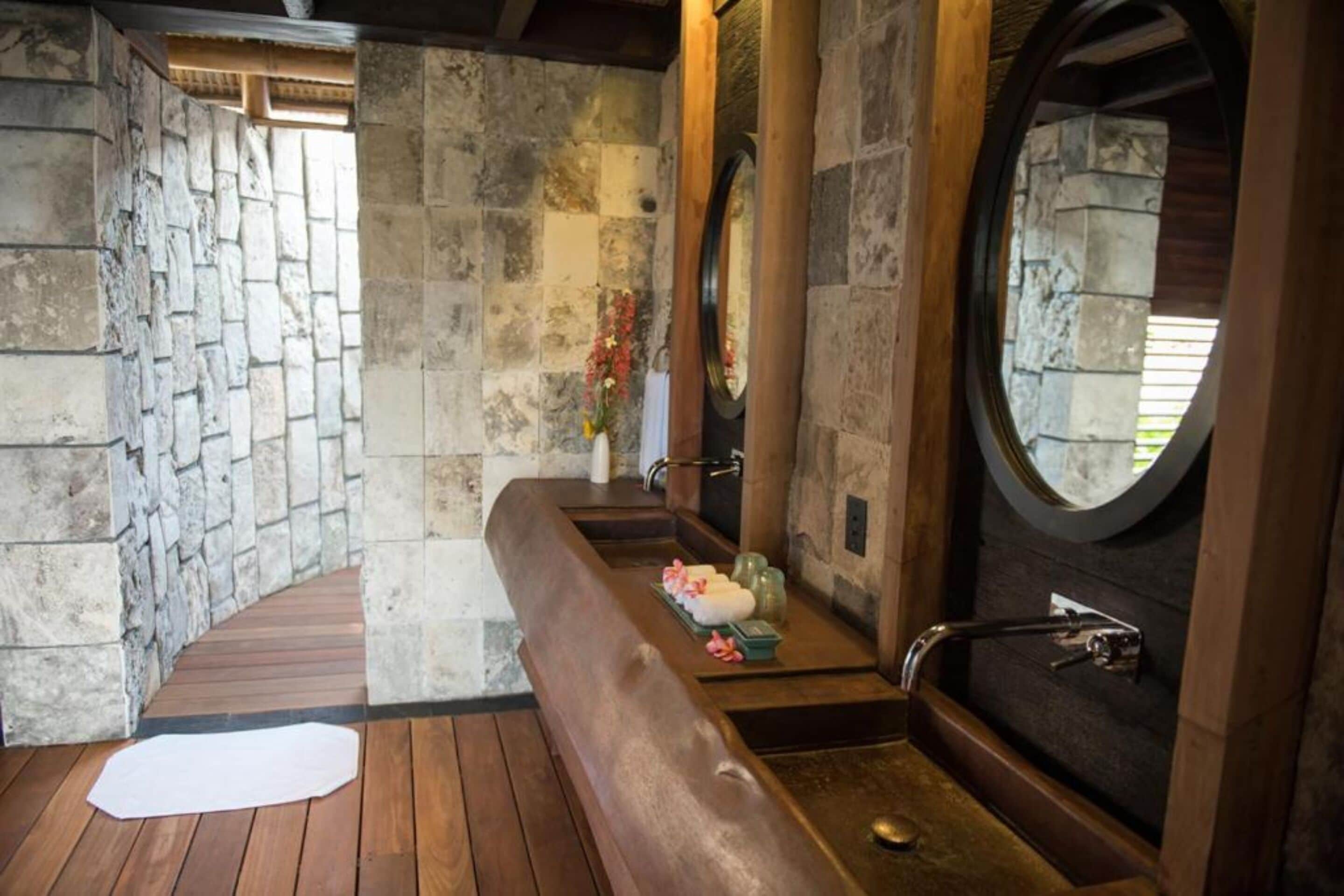 A stone-walled bathroom with a wooden bench, a large round mirror, and a bathtub.