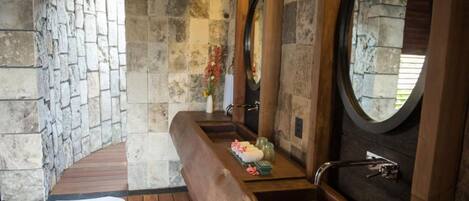 A stone-walled bathroom with a wooden bench, a large round mirror, and a bathtub.