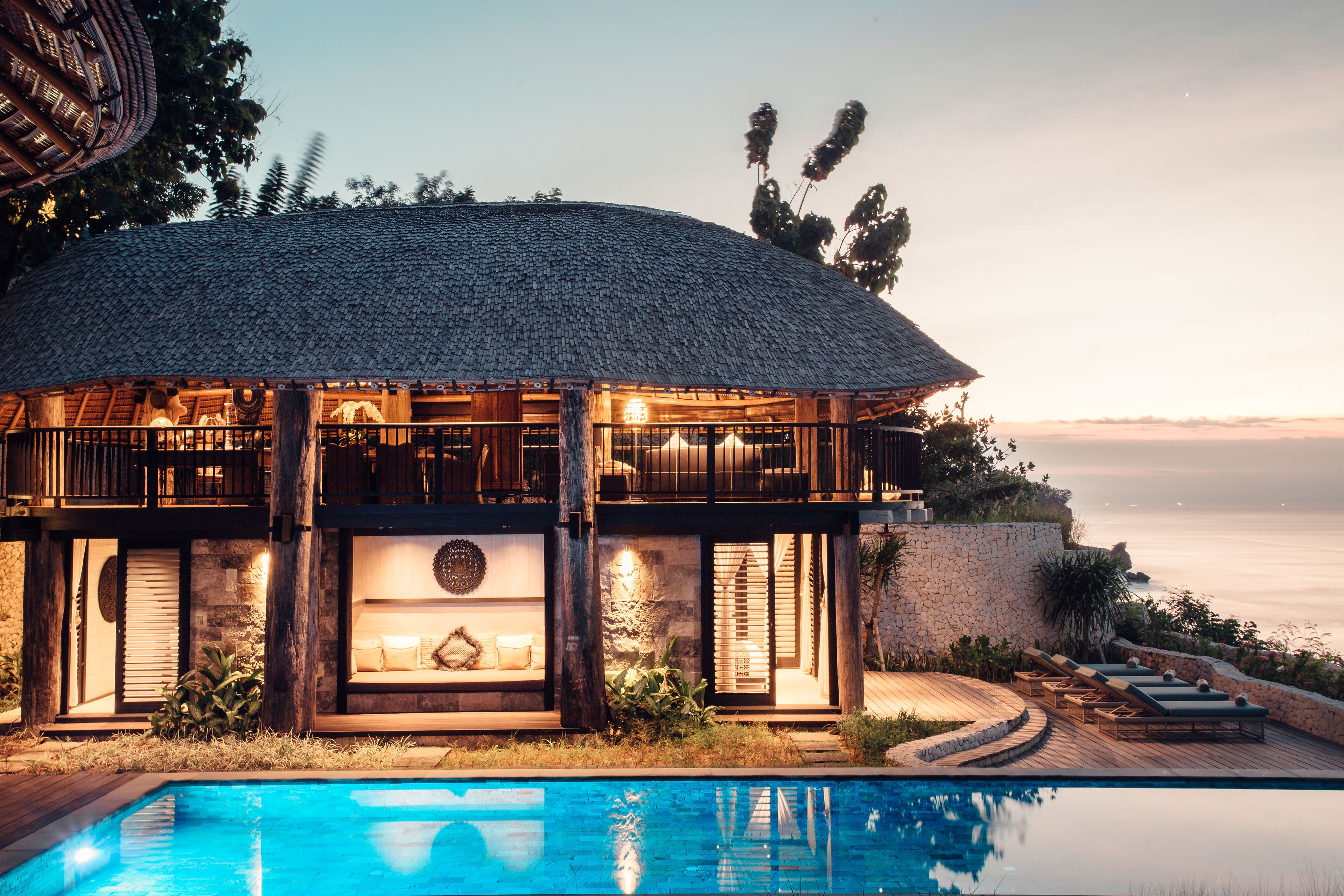 A luxurious villa with a large swimming pool, a thatched-roof structure, and a view of the sea.